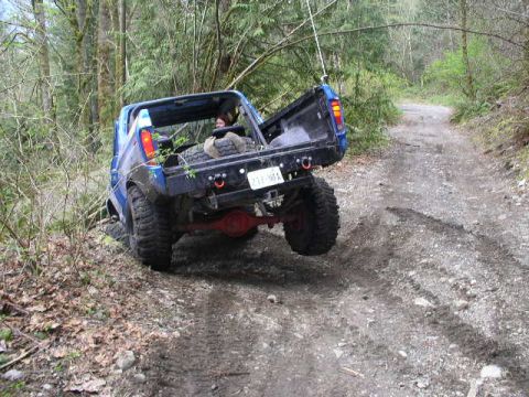 1979 Ford Bronco - A blue off-road vehicle navigates a wooded trail with its tailgate open.