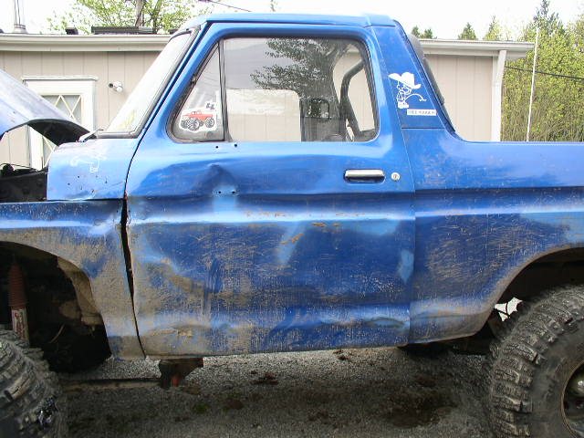 Side of a blue dent-cab Ford truck with sun-faded paint and lift, monster-truck decal in window