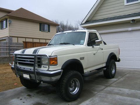 1991 Ford Bronco - Front three-quarter driver-side view of a lifted white Ford Bronco with black hood stripes, chrome tube grille guard, chrome tow mirrors, b…