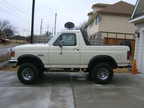 1991 Ford Bronco - Passenger-side profile of a lifted white full-size Ford Bronco with the hardtop off and soft-top bows folded down, sitting on a concrete dr…