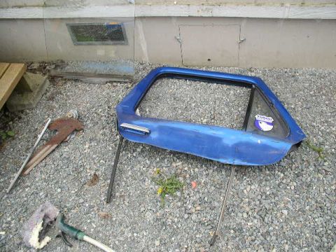 1979 Ford Bronco - A blue door, likely from a vehicle, rests on a gravel surface near a building.