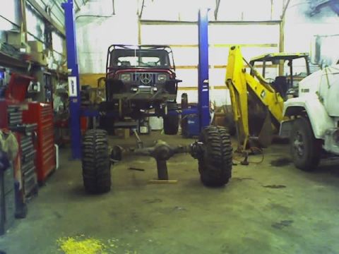 1992 Ford Bronco - A Jeep CJ-7 is suspended on a lift in a workshop, with heavy machinery nearby.