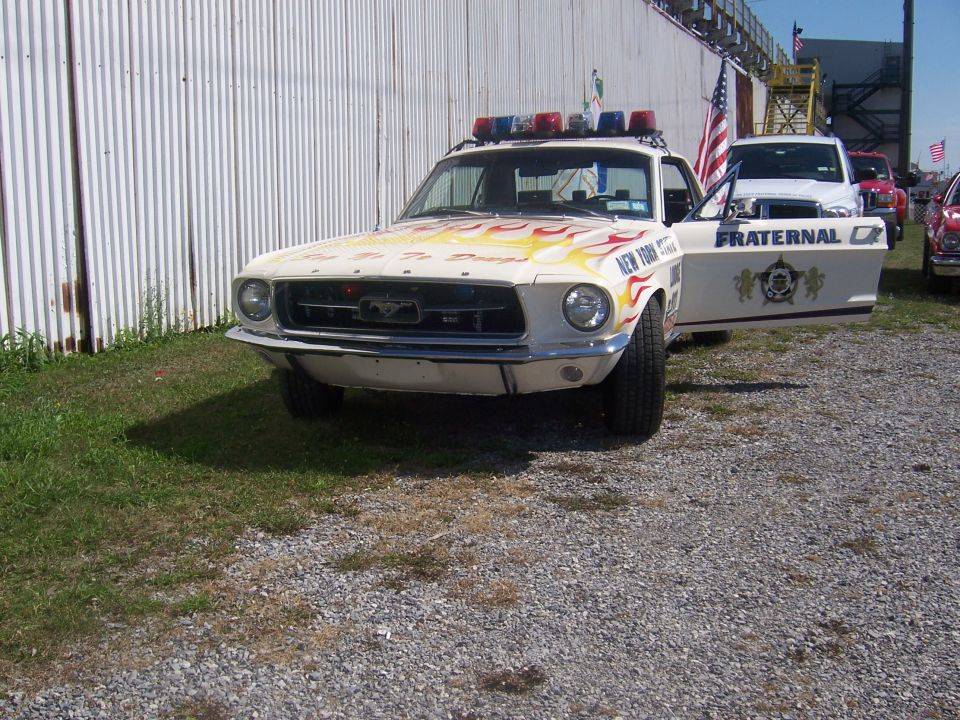 White police car with flame decals and light bar, front view. Appears to be a 1967 Ford Mustang