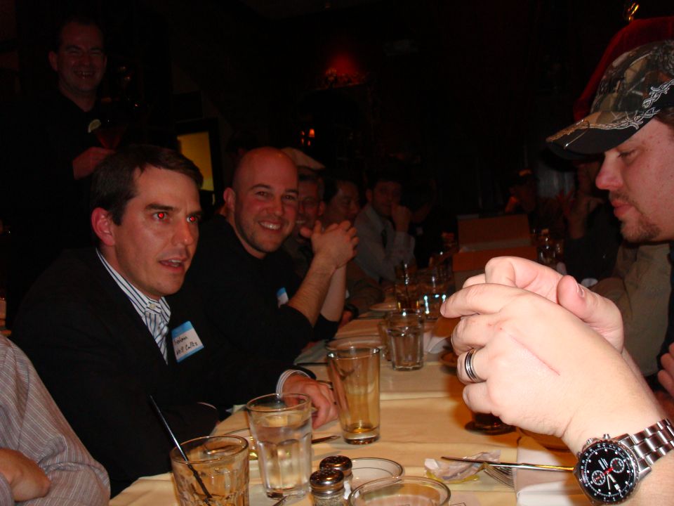 Several men are seated at a table, some smiling, with drinks and place settings