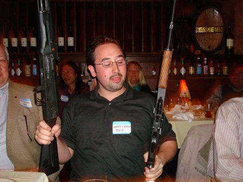 1990 Ford Bronco - A man holds two rifles while seated at a table with others in a dimly lit room.