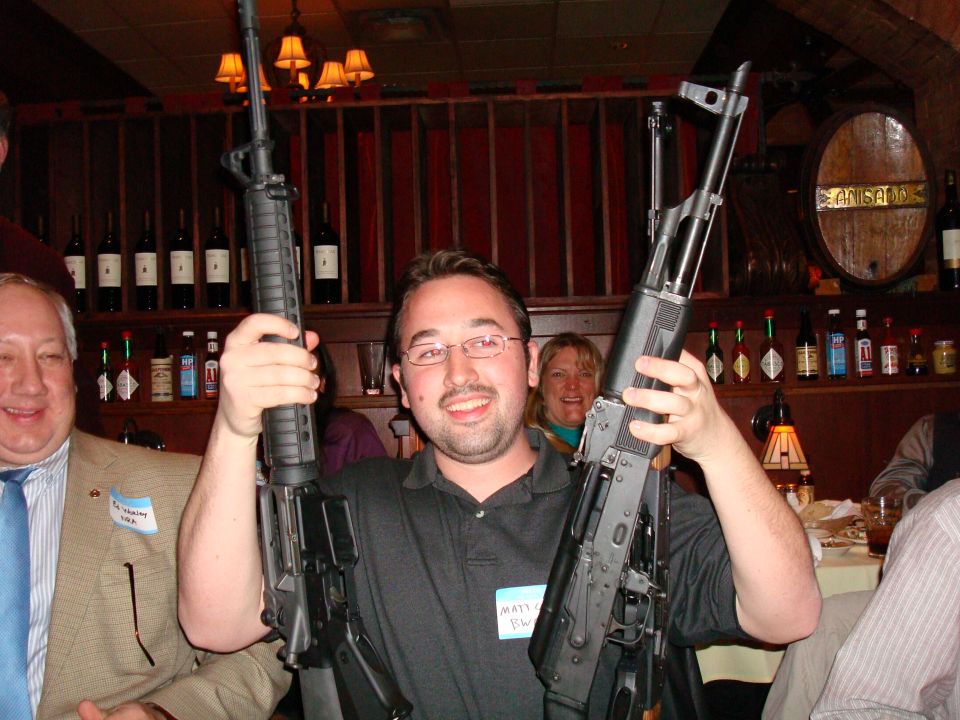guy in black shirt holding AK-47, guy in tan jacket holding AR-15, people in background