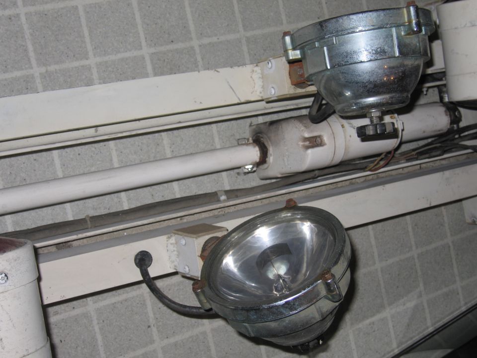 Two industrial spotlights with metal housings and glass lenses are mounted on a white metal track against a tiled wall