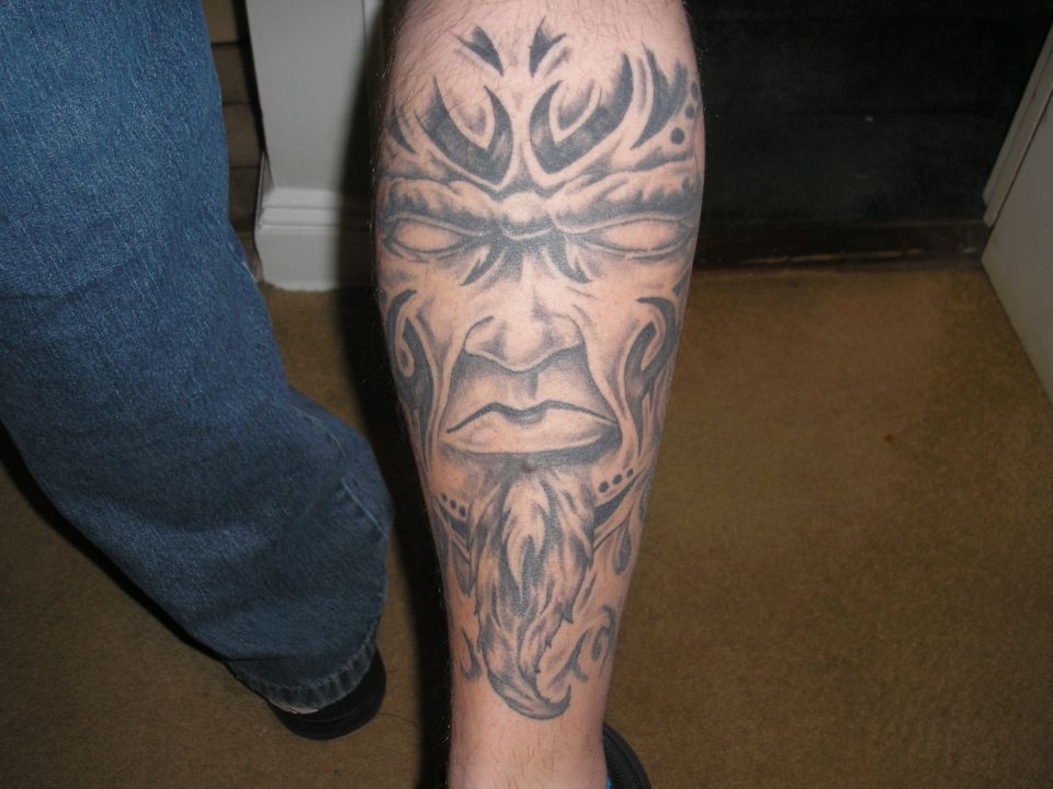 Tattoo of a tribal face with a beard on a leg, next to blue jeans