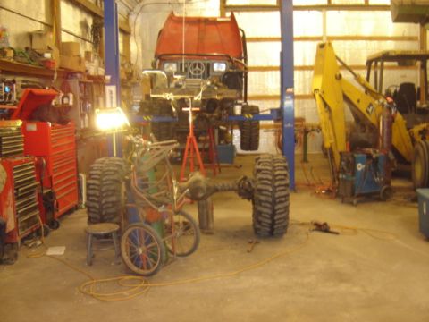 1992 Ford Bronco - A red truck chassis is elevated on a lift in a workshop, with a yellow backhoe and various tools visible in the background.