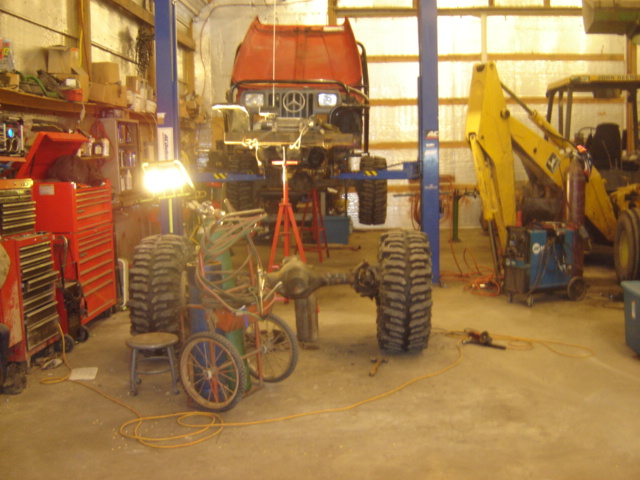 Red truck chassis on lift in garage with yellow backhoe and tools