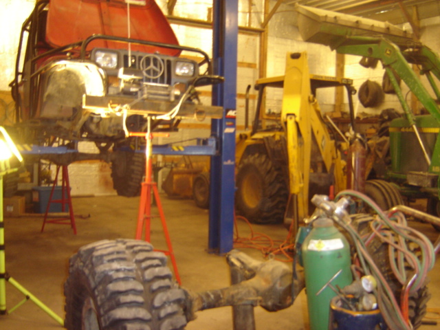 Jeep Wrangler on a lift, front end visible. Yellow backhoe and green tractor in background