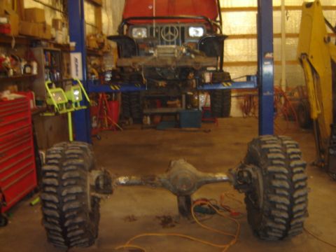 1992 Ford Bronco - A red Mercedes-Benz G-Wagen is suspended on a lift, with its front end visible and a detached axle and large tires in the foreground.