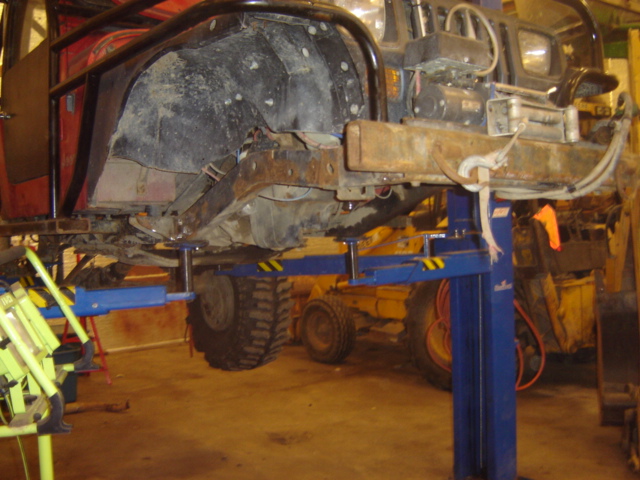 Off-road vehicle chassis on a lift, showing the underside and suspension components. A yellow piece of heavy equipment is visible in the background