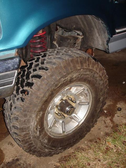 1996 Ford F-150 - Close-up of a teal truck's front wheel and suspension system, showing a red coilover shock and a muddy tire on a silver alloy rim.