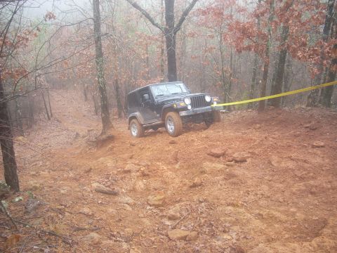 1992 Ford Bronco - A black Jeep Wrangler navigates a steep, muddy incline in a wooded area, with a yellow tow strap attached.