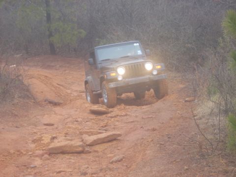 1992 Ford Bronco - A Jeep Wrangler navigates a challenging off-road path with muddy terrain and scattered rocks.