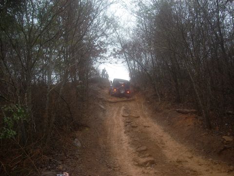 1992 Ford Bronco - A dark Jeep Wrangler navigates a challenging muddy trail, with other vehicles and people visible further up the path.