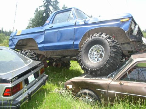 1979 Ford Bronco - A heavily modified blue truck is precariously perched atop two other vehicles in a field.