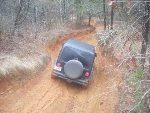 1992 Ford Bronco - A black Jeep Wrangler tackles a challenging muddy trail surrounded by trees.