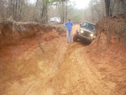 1992 Ford Bronco - A dark Jeep Wrangler navigates a challenging muddy off-road trail, with another vehicle visible in the background.