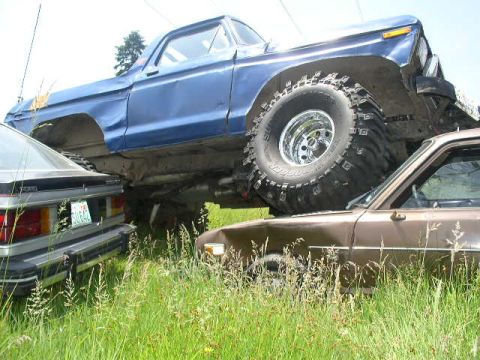 1979 Ford Bronco - A heavily modified blue Ford Bronco from 1978 is parked on top of a brown car in a grassy field.