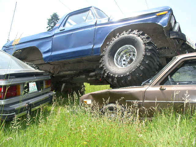Blue Ford 4x4 on huge Super Swamper tires straddling two crushed cars in tall grass
