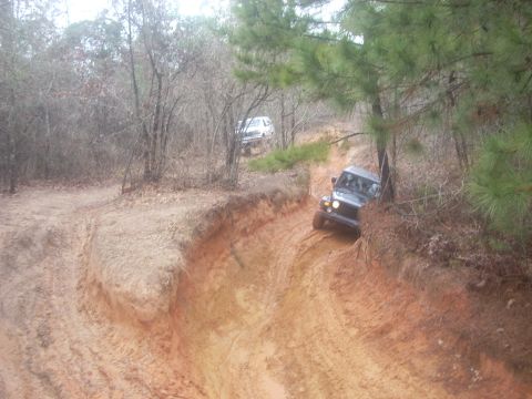 1992 Ford Bronco - Two off-road vehicles navigate a challenging muddy track through a wooded area.