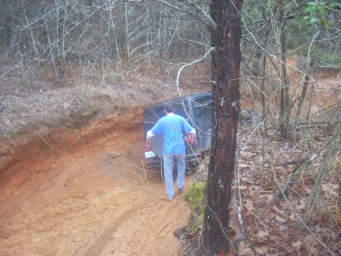 1992 Ford Bronco - A person walks towards a black SUV that is stuck in a muddy, wooded area. The rear of the vehicle is visible.
