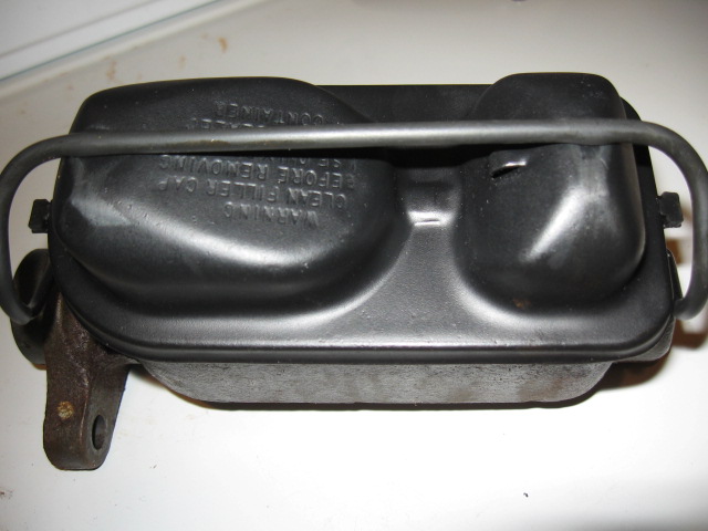 Black brake master cylinder with metal mounting bracket. Text on top reads 'WARNING CLEAN FILLER CAP BEFORE REMOVING CONTAINER'