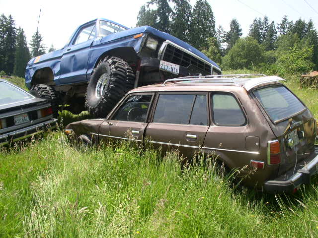 Blue lifted Ford 4x4 crushing a brown Honda station wagon in a field
