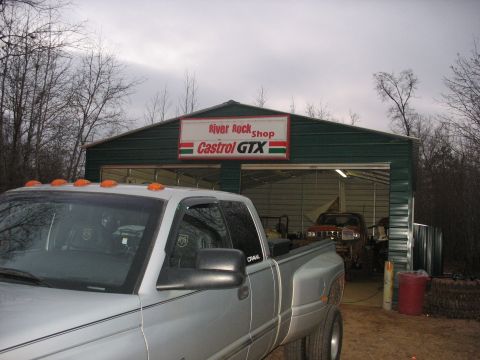 1978 Ford Bronco - A silver pickup truck is parked outside a garage with a Castrol GTX sign, while another vehicle is visible inside.