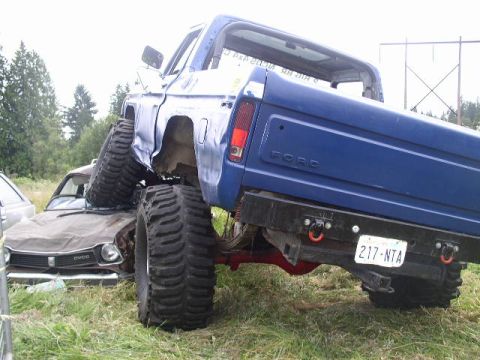 1979 Ford Bronco - A blue 1979 Ford Bronco is parked on top of a brown car, with its large tires crushing the vehicle below.