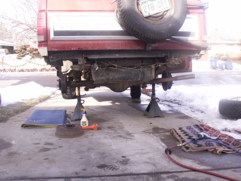 1986 Ford Bronco - The rear of a red SUV is elevated on jack stands for maintenance, revealing its suspension and exhaust system.