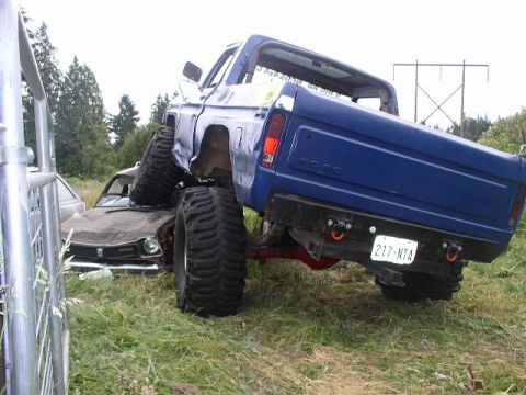 1979 Ford Bronco - A heavily modified blue pickup truck is shown resting on top of a car, with its large tires dominating the scene.