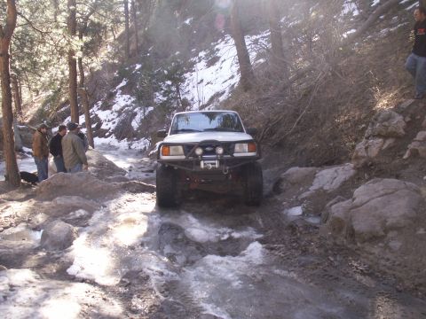 1986 Ford Bronco - An off-road vehicle carefully makes its way through a challenging, icy, and muddy trail surrounded by trees.