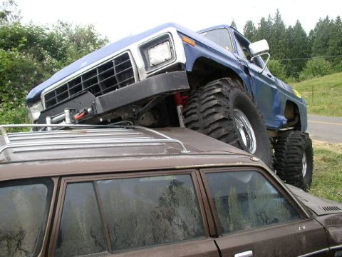 1979 Ford Bronco - A lifted blue truck is precariously balanced on top of a brown station wagon.