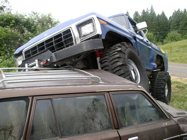 Blue and white Ford Bronco on mud tires crushing an old brown station wagon