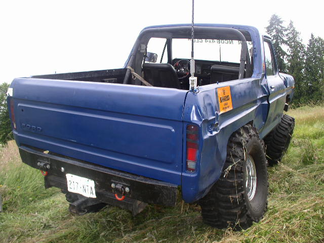 Blue pickup truck with large tires, rear view. The vehicle has a dent on the rear quarter panel and a yellow sticker on the side