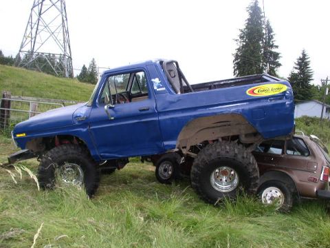 1979 Ford Bronco - A heavily modified blue truck is precariously balanced on a smaller brown car in an outdoor setting.