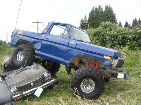 1979 Ford Bronco - A blue 1979 Ford Bronco is perched atop a severely damaged car, highlighting its off-road capabilities.