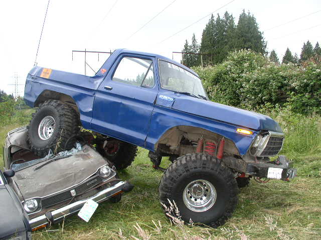 Lifted blue Ford Bronco on mud tires climbing over a crushed gray compact car