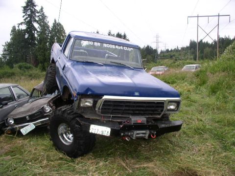 1979 Ford Bronco - A modified blue pickup truck is parked on top of a wrecked car in a grassy field.