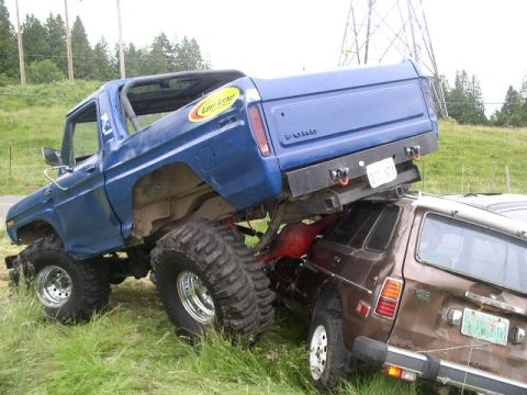 1979 Ford Bronco - A lifted blue 1979 Ford Bronco is positioned on top of a brown car, with its large tires resting on the smaller vehicle.