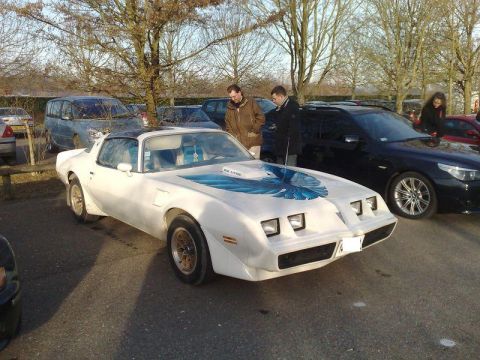 1991 Ford Crown Victoria - A white 1977 Pontiac Firebird Trans Am with a blue hood decal is parked in a lot with other vehicles and people nearby.