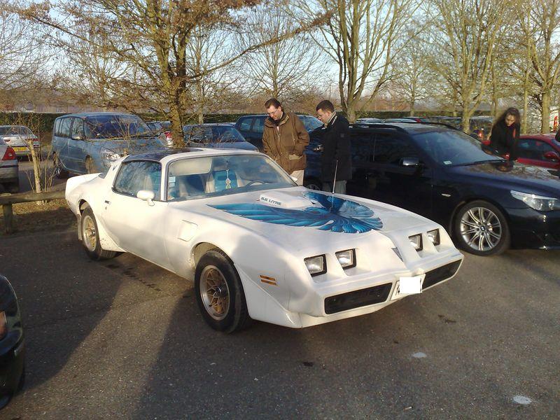 White Pontiac Firebird Trans Am with blue hood decal parked outside, people looking at it