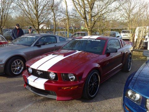 1991 Ford Crown Victoria - A red Ford Mustang with white racing stripes is parked next to other vehicles at an outdoor car show.