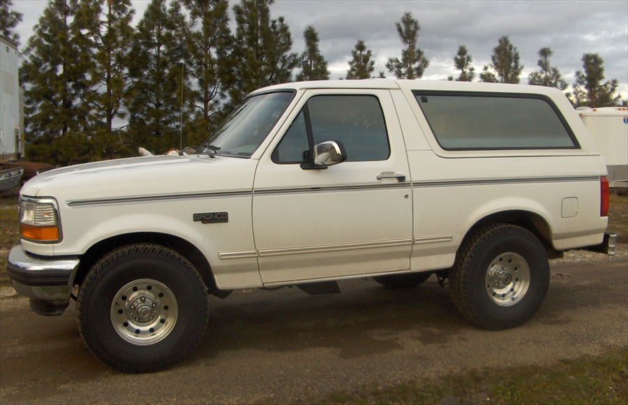 White Ford Bronco SUV in side profile with alloy wheels, parked on gravel with pine trees behind