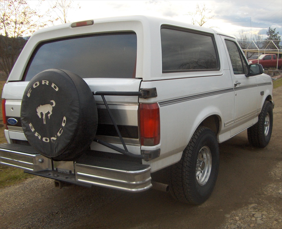 White Ford Bronco SUV rear view with black FORD BRONCO spare tire cover on tailgate, parked on gravel