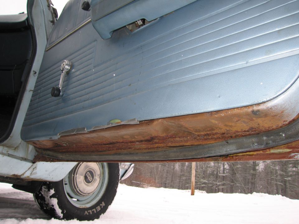interior of a car door with blue paneling, manual window crank, and significant rust on the rocker panel below