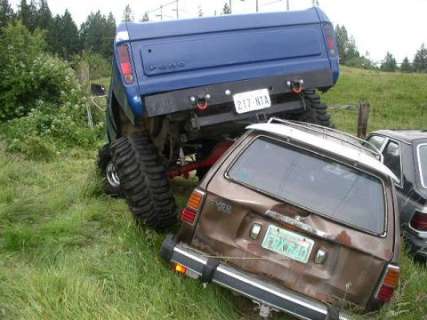 1979 Ford Bronco - A blue 1979 Ford Bronco is precariously perched atop a brown station wagon in a grassy field.